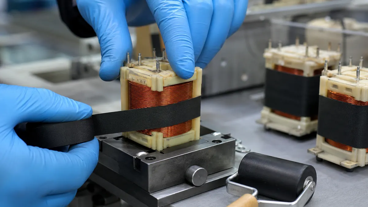 Technician wrapping matte black acetate cloth tape around a wound relay coil bobbin at an assembly fixture